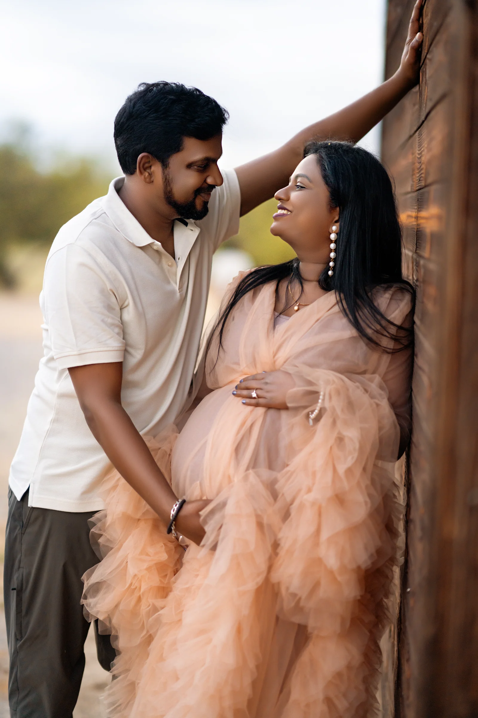 Expectant mother in a peach gown smiling at her partner beside a wooden wall