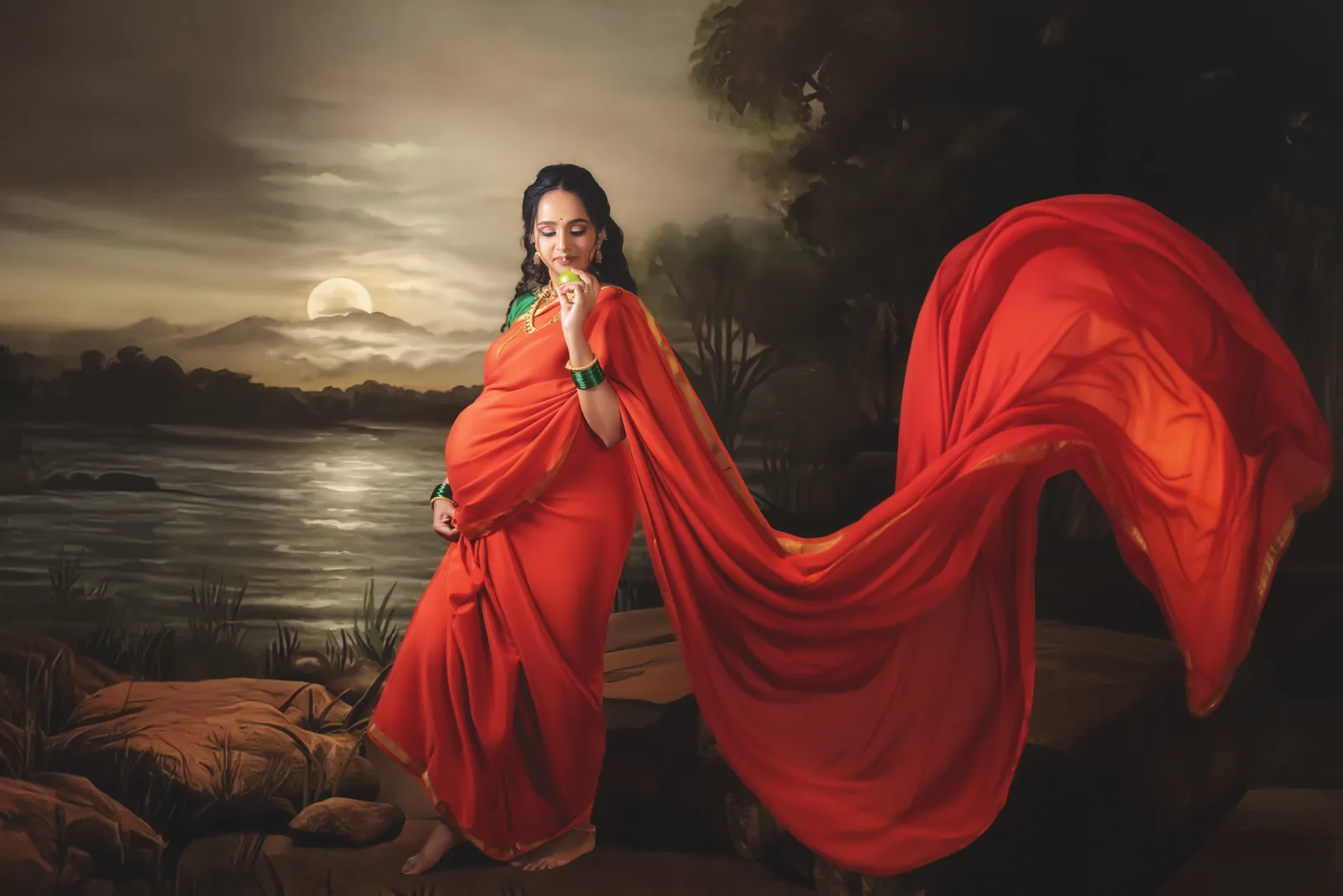 Expectant mother in a red sari with fabric flowing across a dramatic lakeside scene