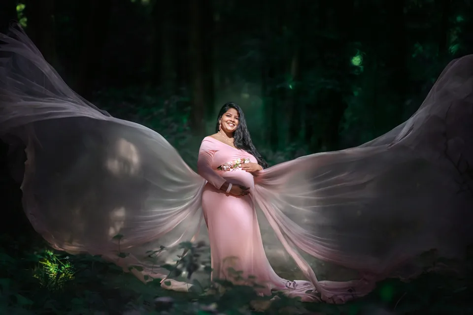 Expectant mother in a pink gown with flowing fabric against a dark woodland background