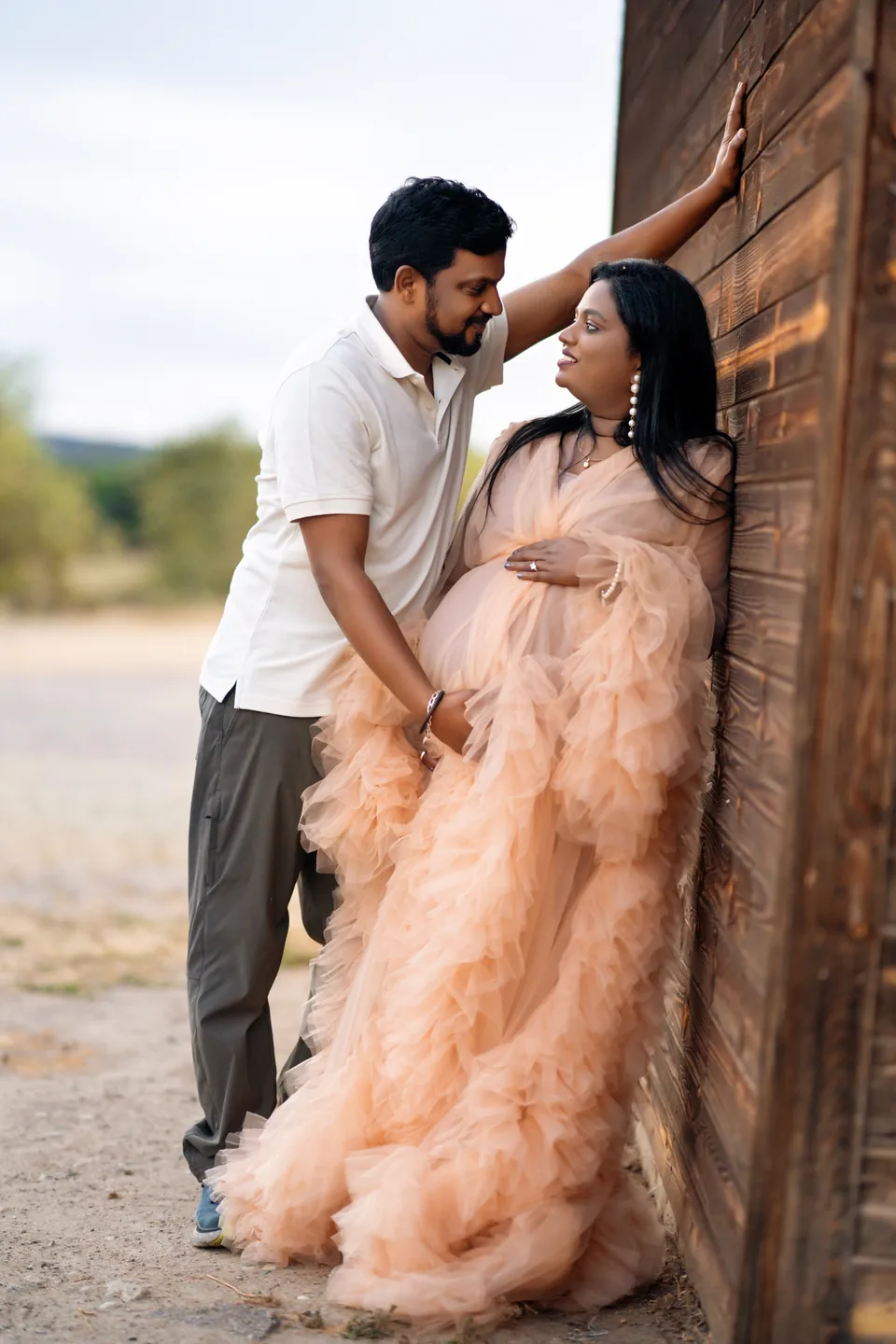 Expectant mother in a peach gown leaning toward her partner beside a wooden wall