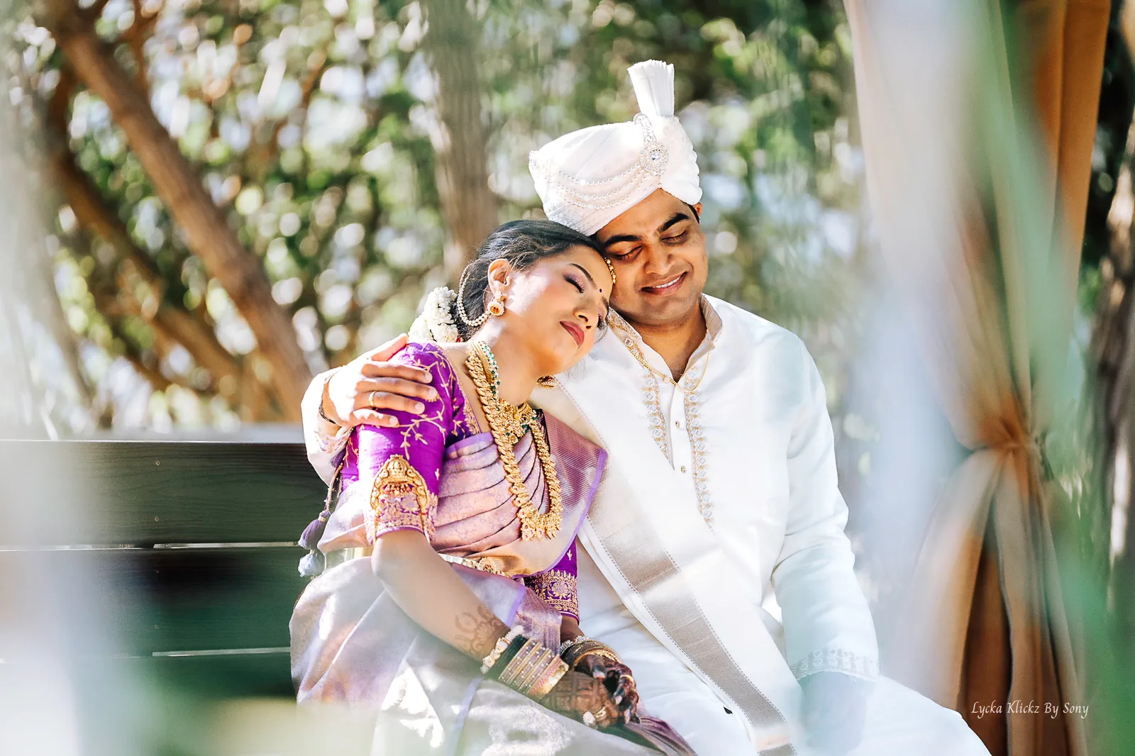 Couple in traditional wedding attire leaning together beneath trees